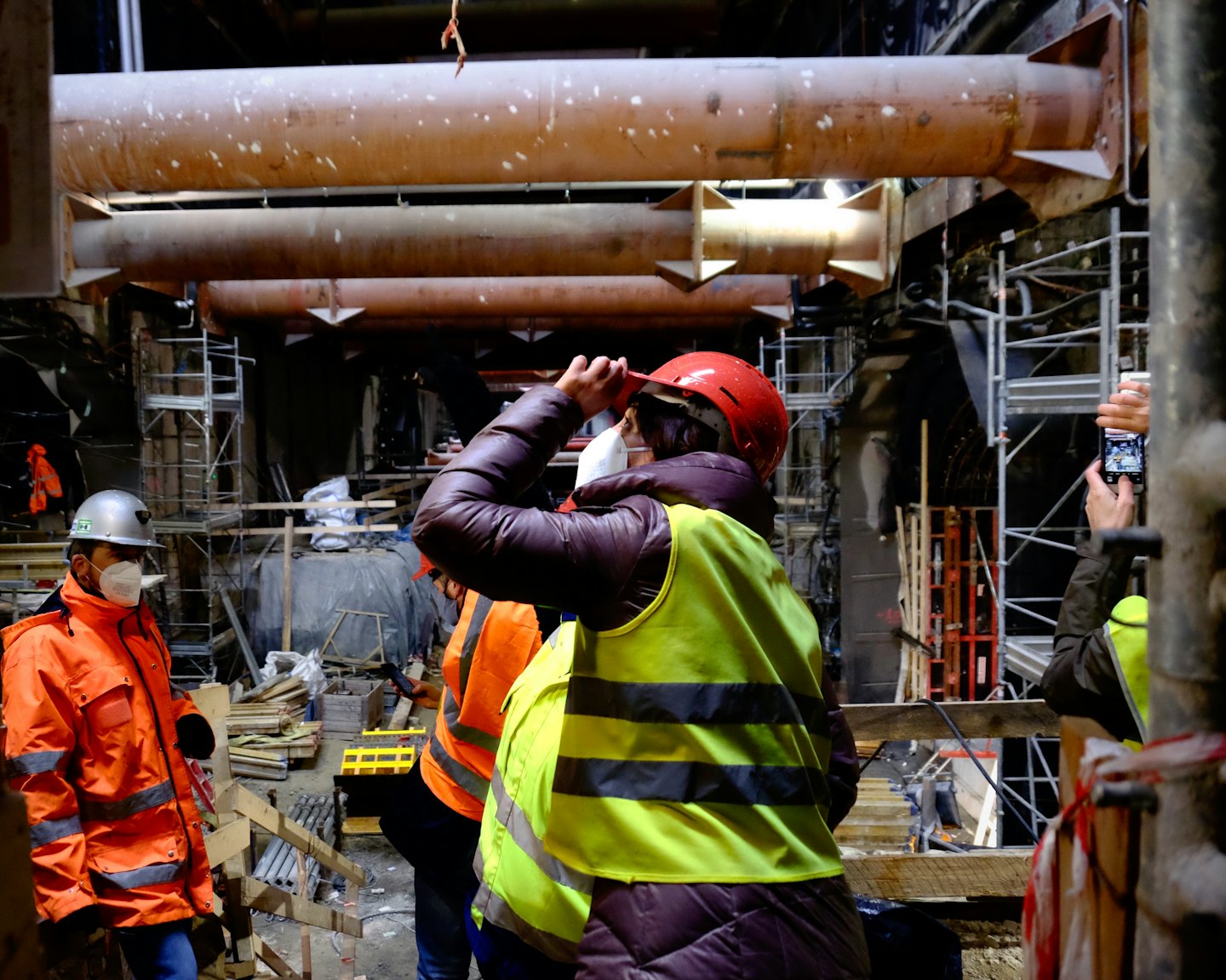 Construction workers examine a tunnel with large pipes and scaffolding.